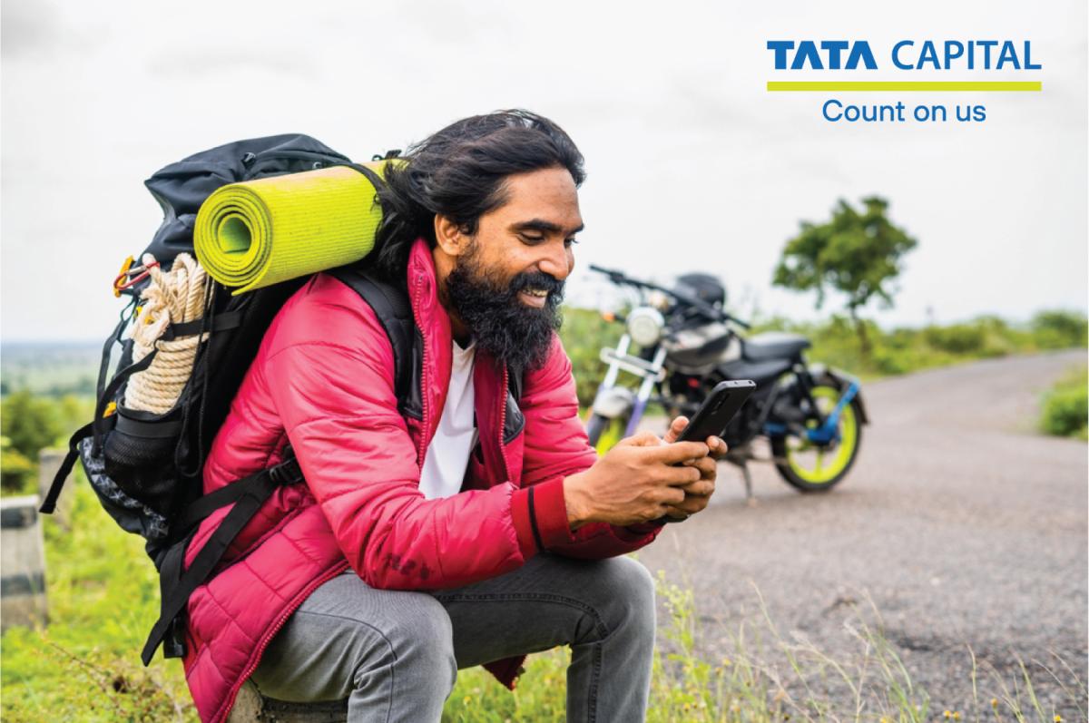 Tata Capital's tips to reduce insurance premiums while a man wearing a red jacket and a backpack is sitting on a rock and using his smartphone.