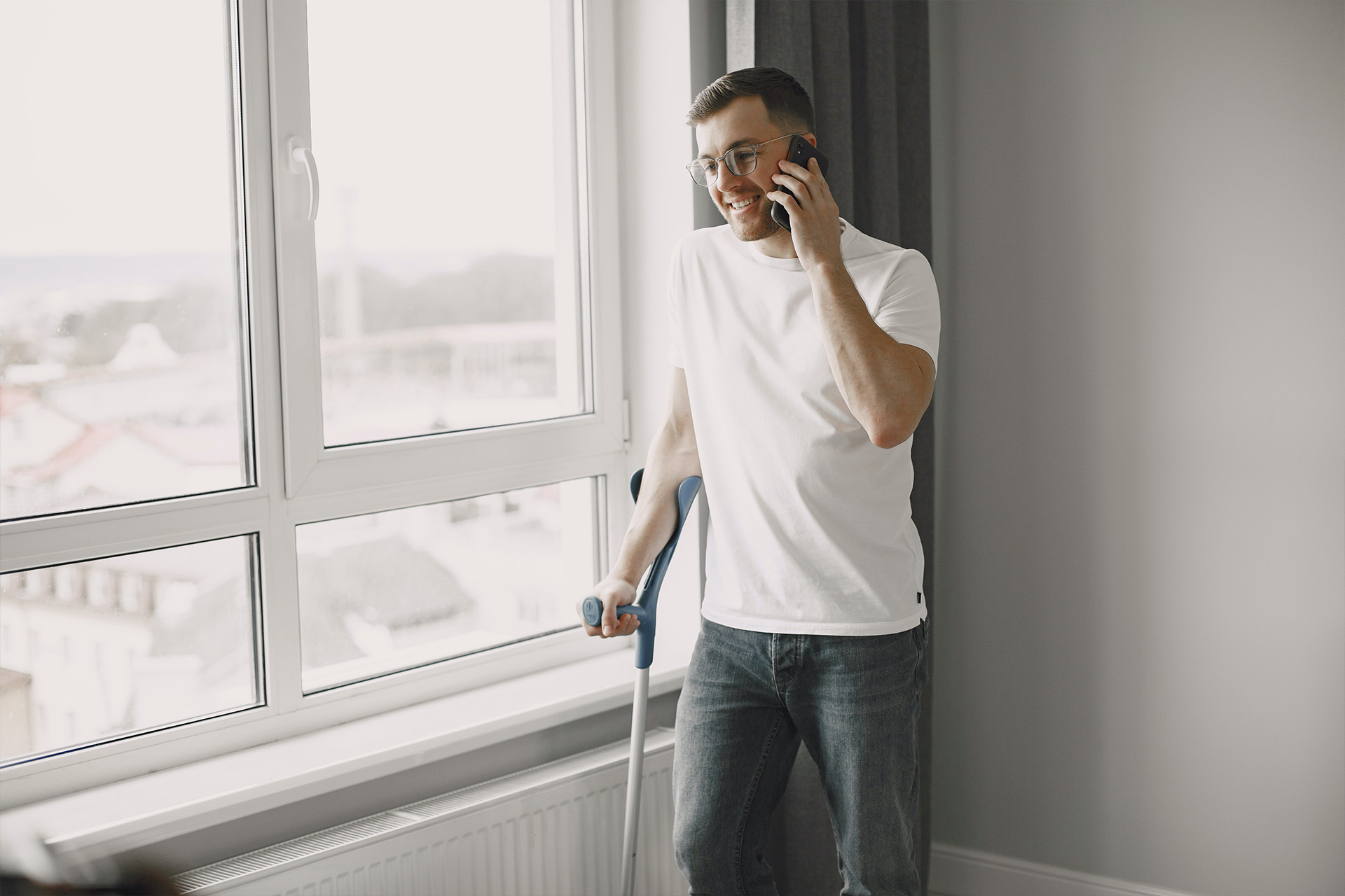 A man with glasses and a cast on his leg is talking on the phone while standing in front of a window.