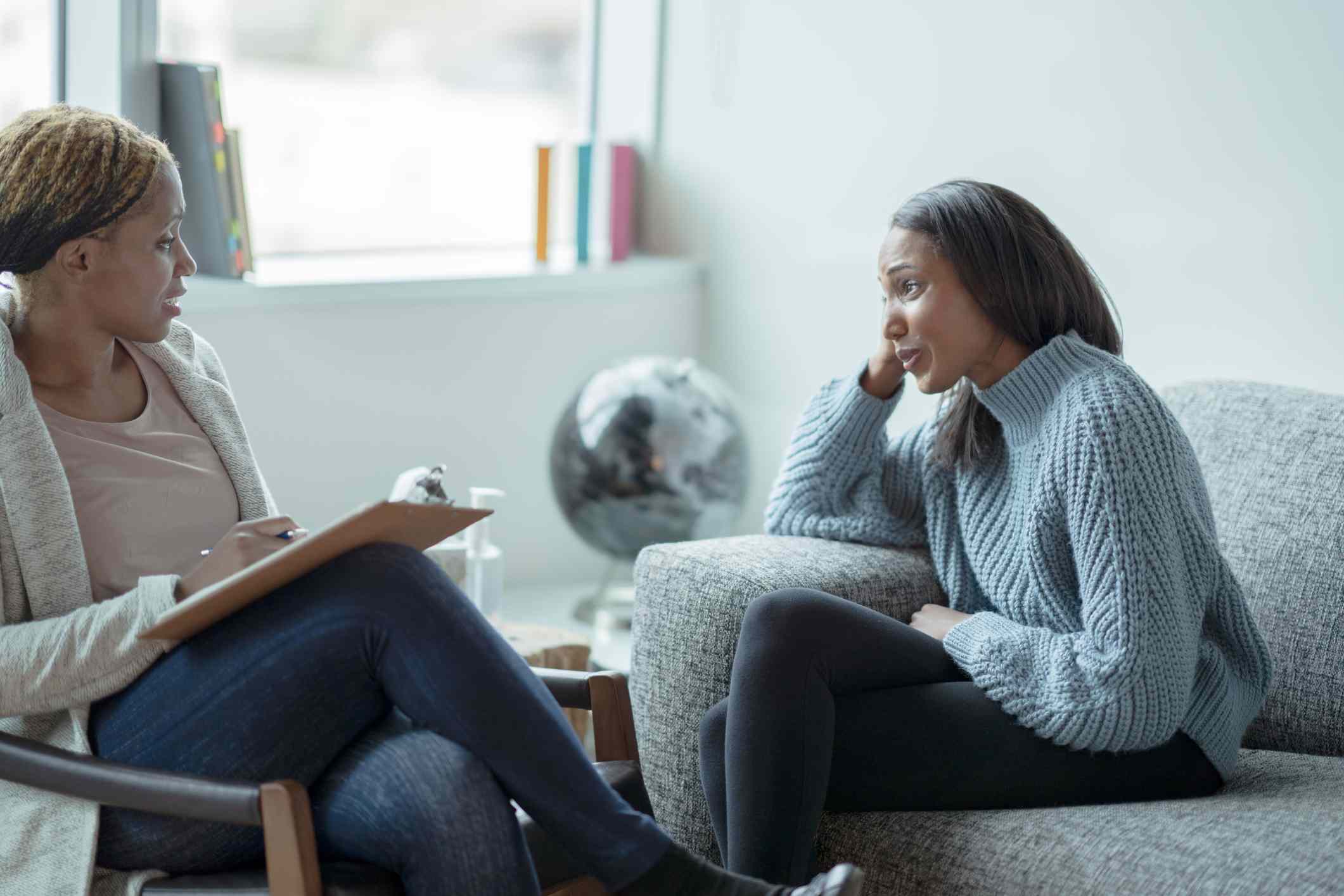 A therapist sits in a chair across from a patient who is sitting on a couch in a clinical setting.